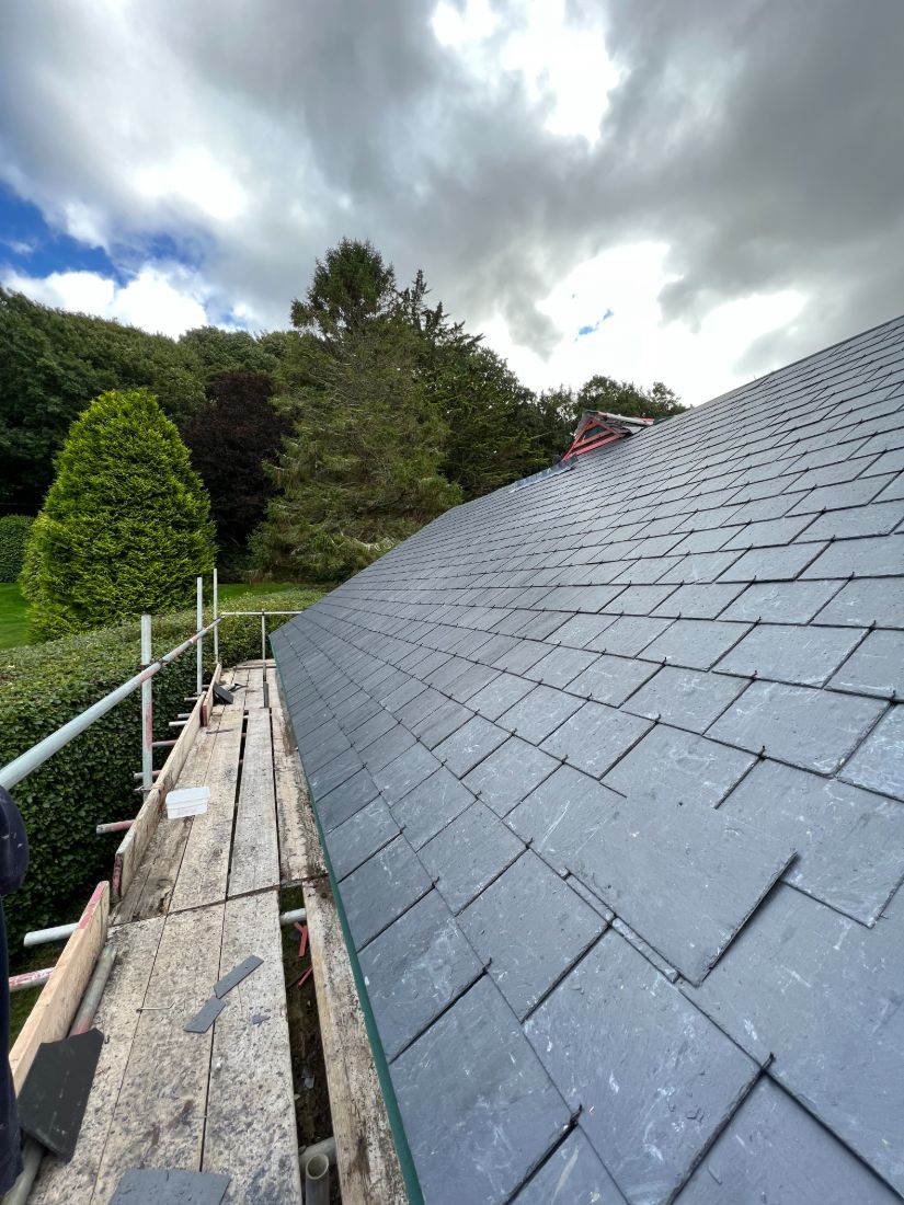 A pitched roof re-laid in natural slate, photographed from the scaffold with a wooded Cornish valley behind